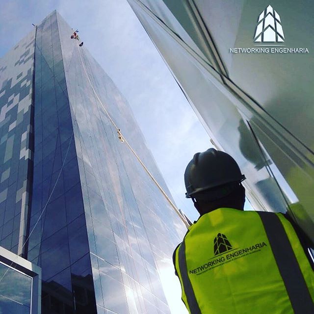Manutenção de Fachadas com Alpinismo Industrial no Rio de Janeiro Técnico da Networking Engenharia, uniformizado com colete e capacete, observa um alpinista industrial realizando a manutenção na fachada de vidro de um arranha-céu.