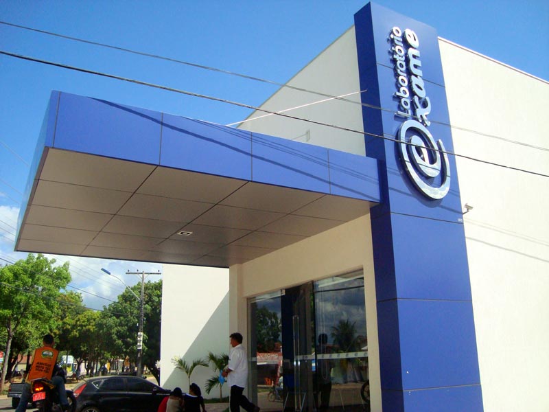 Fachada do Laboratório Exame com uma marquise e um totem vertical revestidos em painéis de ACM azul, com porta e janela de vidro na entrada.