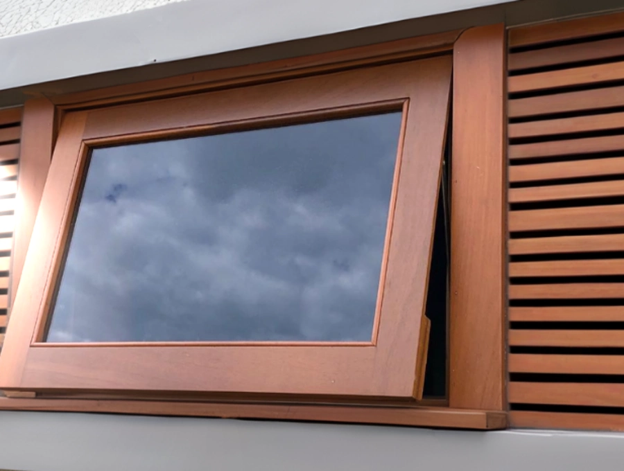 Mecanismo de abertura de uma janela do tipo basculante em madeira, ideal para controlar a ventilação de ambientes como cozinhas e banheiros. Close-up de uma janela basculante com esquadria de madeira em tom de mel, com o vidro escuro refletindo nuvens e uma veneziana de madeira ao lado.