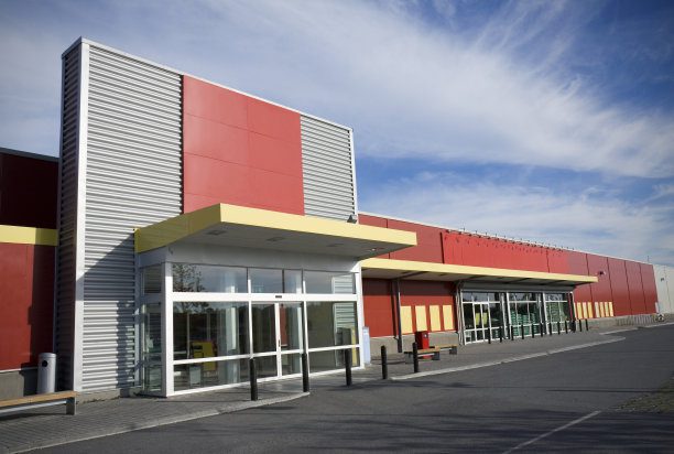 Fachada de um grande estabelecimento comercial, como um supermercado ou loja de departamento, com revestimento que combina painéis de ACM vermelho e amarelo com metal corrugado.