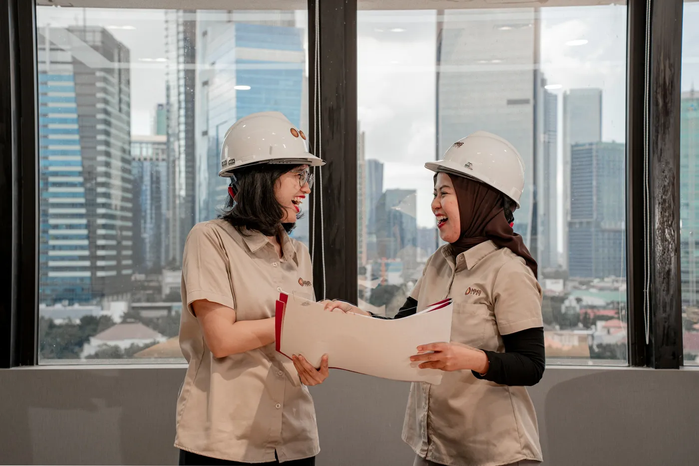 Duas engenheiras de capacete conferem checklist e plantas técnicas em prédio comercial com vista para o centro da cidade
