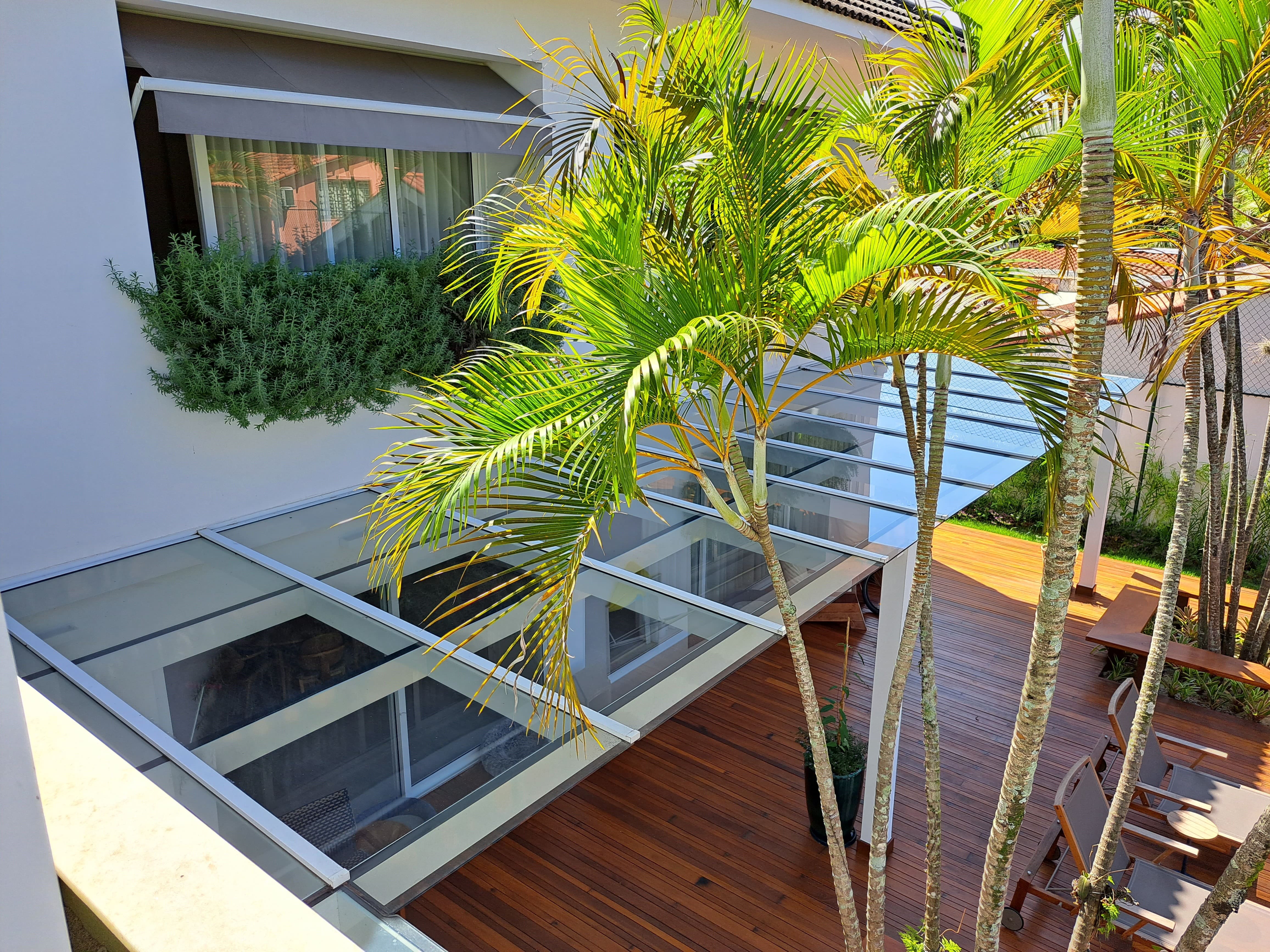 Cobertura de pergolado de vidro com estrutura de alumínio branco, instalada sobre um deck de madeira em uma área de lazer moderna com palmeiras.