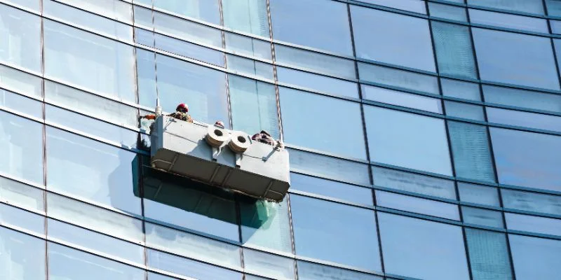 Equipe de limpeza profissional em um andaime suspenso (balancim) realizando a manutenção e limpeza da fachada de vidro de um grande edifício.