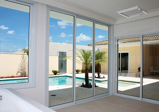 Porta de Vidro para Piscina: Integre seus Ambientes com Estilo Quarto com grandes portas de correr de vidro e esquadrias de alumínio branco, com vista para uma área de lazer com piscina e um céu azul ensolarado.