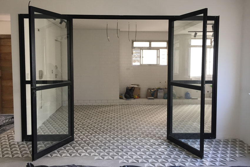 Porta de Vidro Estilo Industrial: Serralheria Preta para Divisórias Porta de giro dupla em vidro com serralheria preta em estilo industrial, instalada em um ambiente com piso de ladrilho geométrico e paredes brancas em fase de acabamento.