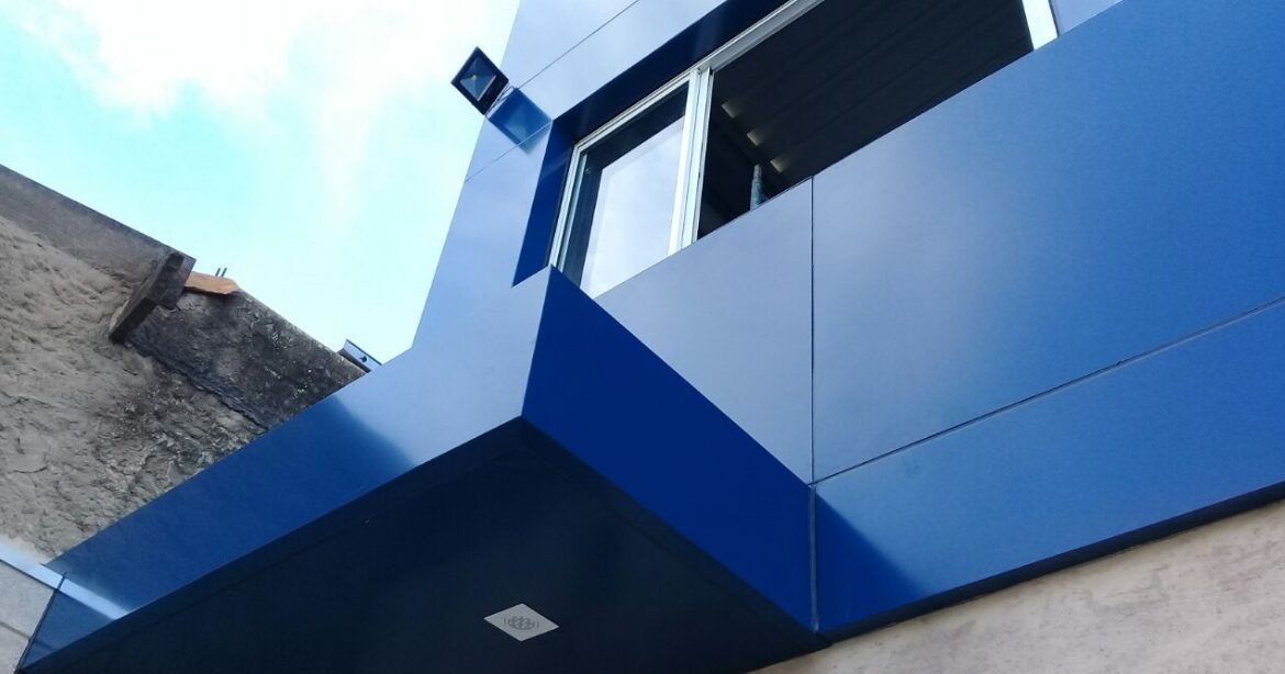 Como Escolher a Espessura do ACM para seu Projeto: Dicas de Engenharia Detalhe de uma fachada com revestimento em painéis de ACM azul, mostrando o acabamento preciso dos cantos e a integração com a janela, ilustrando a aplicação do material na espessura correta.