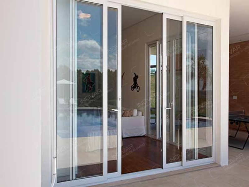 Porta de Correr de 4 Folhas em Alumínio Branco para Salas Porta de correr de quatro folhas com esquadria de alumínio branca e vidro, conectando uma sala de estar com piso de madeira a uma área externa com piscina."