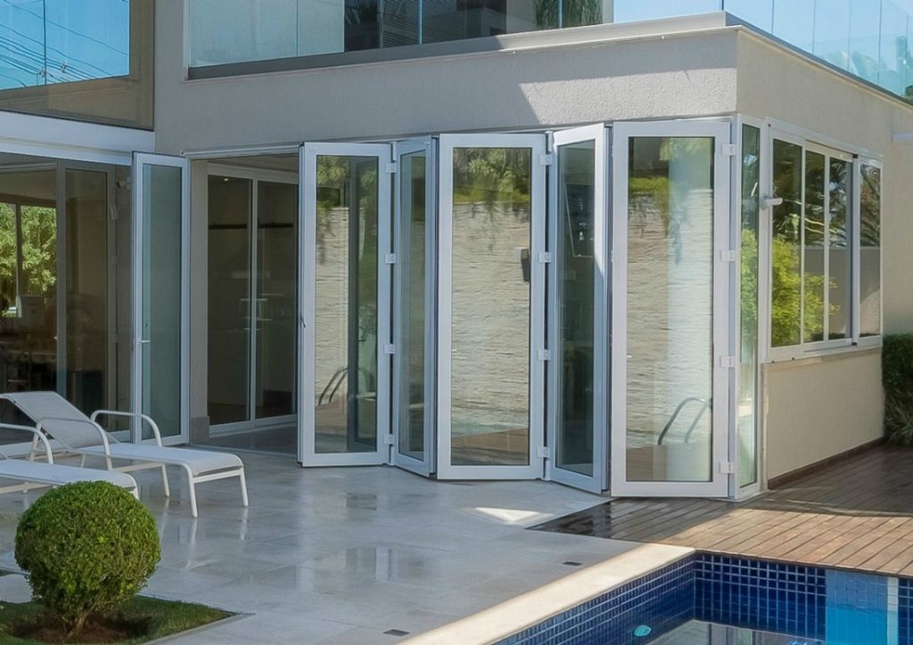 Ampla porta de vidro articulada (camarão) com esquadria de alumínio branco, integrando a casa a uma área de lazer com piscina e deck de madeira.