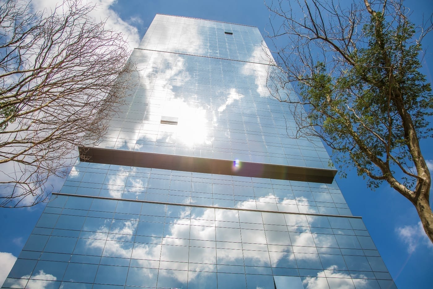 Vista em contraponto da fachada de um arranha-céu com vidro de proteção solar, que reflete intensamente o céu azul e as nuvens, bloqueando o calor.