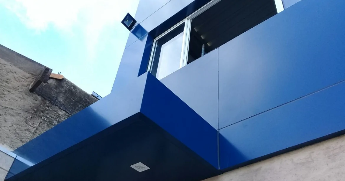 Revestimento em ACM: A Melhor Opção para Reformar sua Fachada Detalhe de uma fachada comercial com revestimento em painéis de ACM azul, mostrando os ngulos precisos, a sanca com iluminação embutida e uma janela de alumínio.