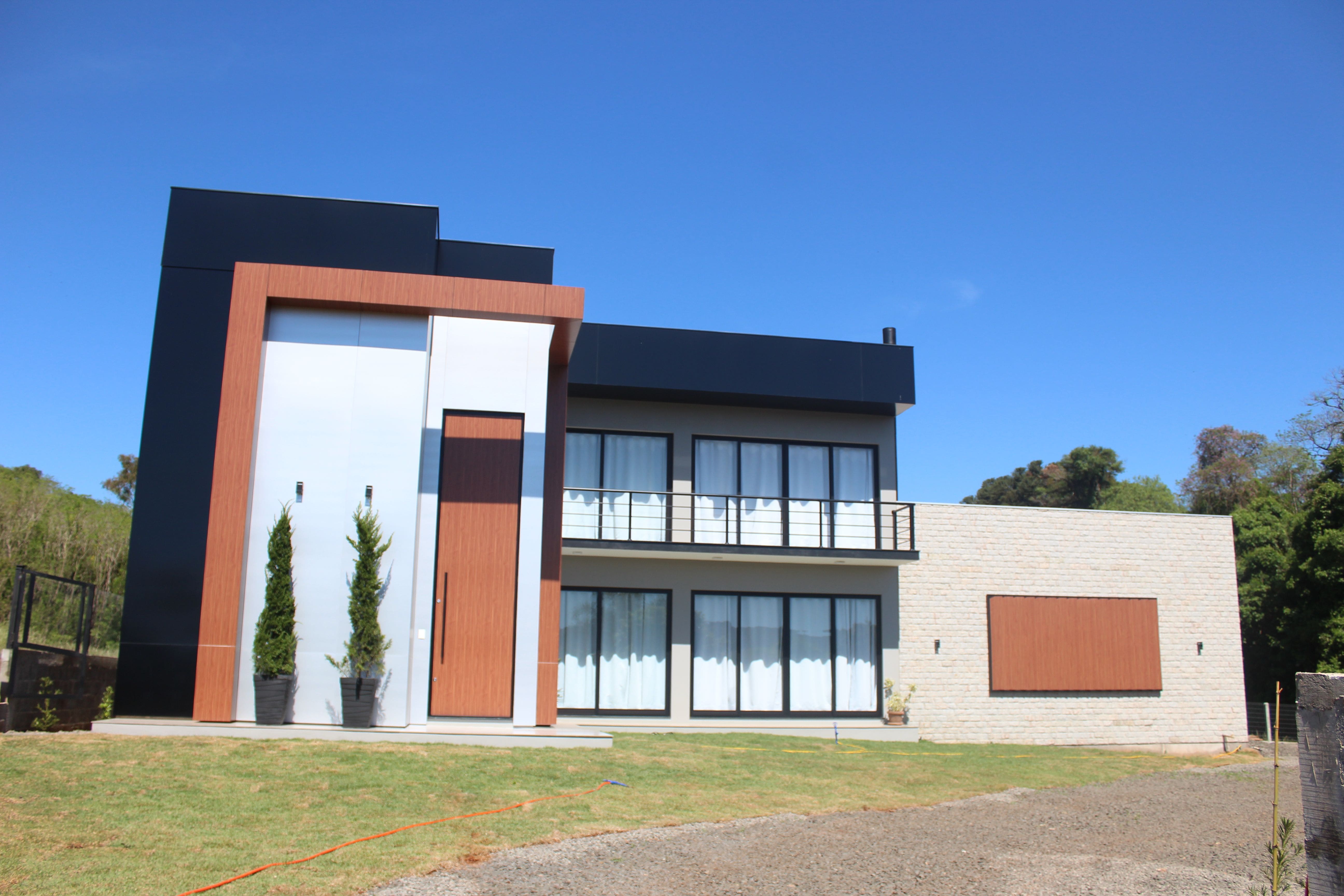 Fachada de um sobrado moderno com revestimento que combina painéis de ACM nas cores preto, prata e amadeirado, além de tijolo aparente e grandes portas de vidro.