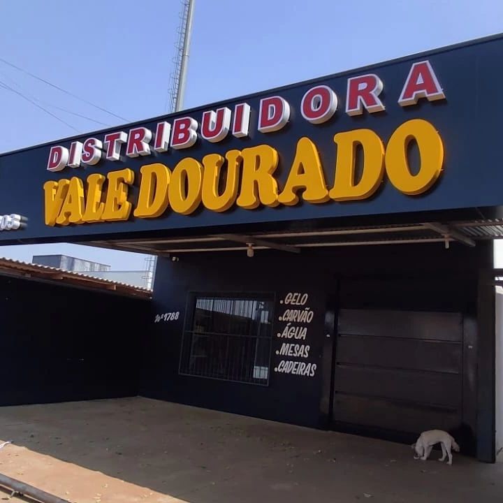 Fachada da 'Distribuidora Vale Dourado', com estrutura preta, letreiro grande em amarelo e vermelho, e uma lista de produtos como gelo e carvão na parede