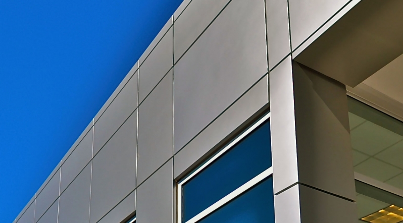 Fachada em ACM Prata: Modernidade e Durabilidade para sua Empresa Close-up da fachada de um edifício revestido com painéis de ACM na cor prata, mostrando o alinhamento das juntas e a integração com uma janela de vidro azulado.