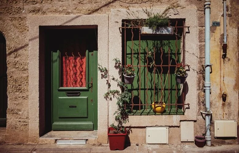 Fachada retrô de uma casa antiga com parede de pedra, uma porta de madeira verde, uma janela com grade de ferro e vasos de plantas.