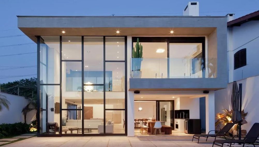 Casa moderna de dois andares com fachada de vidro em pé-direito duplo e esquadrias pretas, iluminada ao anoitecer, com área de lazer e piscina integradas à sala.