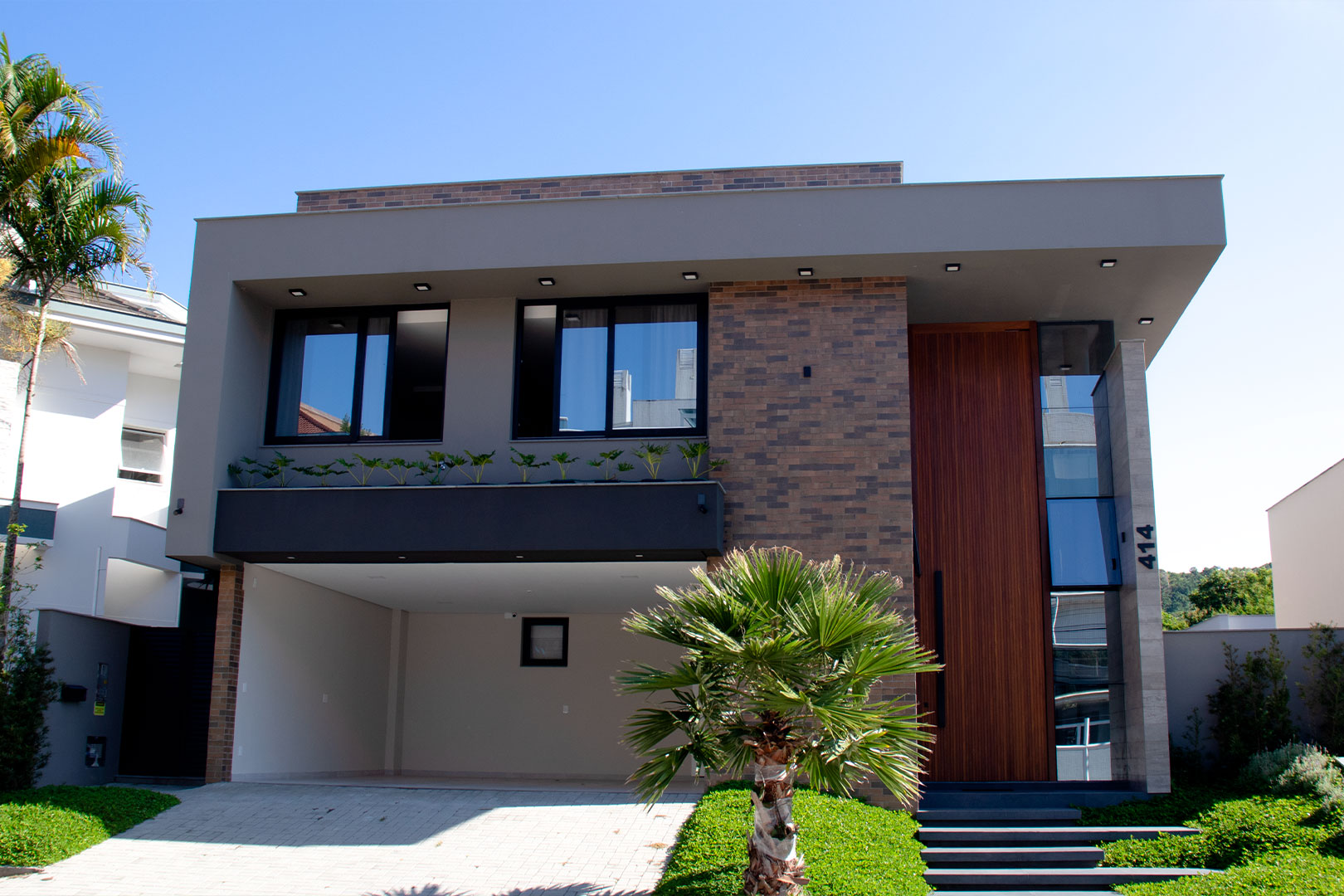 Fachadas Modernas: Como Combinar Vidro (Glazing), Madeira e Pedra em 2025 Fachada de casa moderna de dois andares (sobrado) com pintura cinza, detalhes em tijolinho, porta de entrada imponente de madeira e janelas de vidro com esquadria preta.