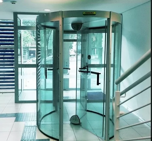 Porta Giratória para Controle de Acesso em Prédios e Condomínios Porta giratória de vidro com estrutura de alumínio instalada na entrada de um edifício comercial, com piso tátil para acessibilidade em frente.