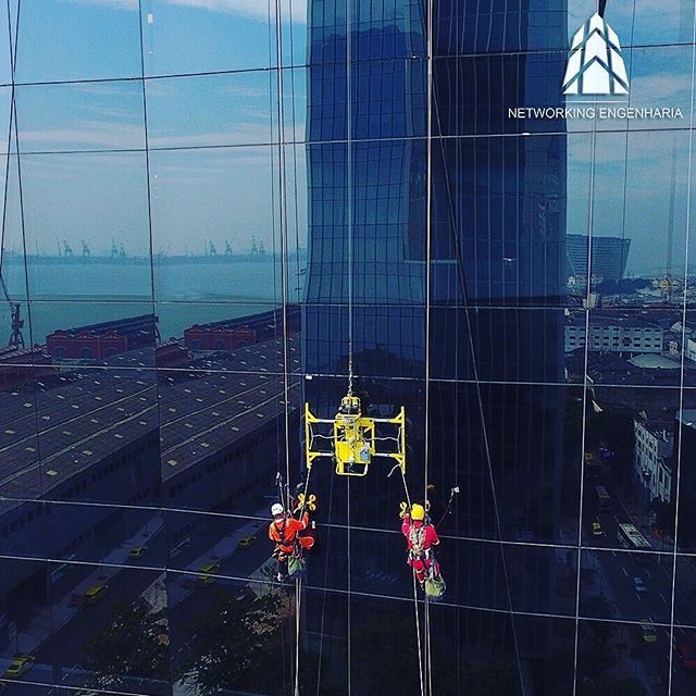 Dois técnicos de alpinismo industrial da Networking Engenharia suspensos por cordas na fachada de vidro de um arranha-céu com vista para o porto do Rio de Janeiro