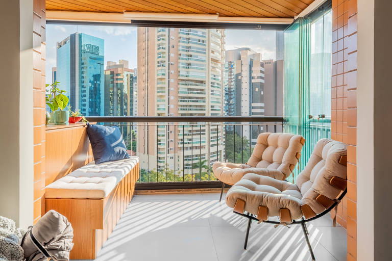 Varanda de apartamento decorada com poltronas confortáveis e fechamento em cortina de vidro, permitindo a vista da cidade com proteção contra o vento.