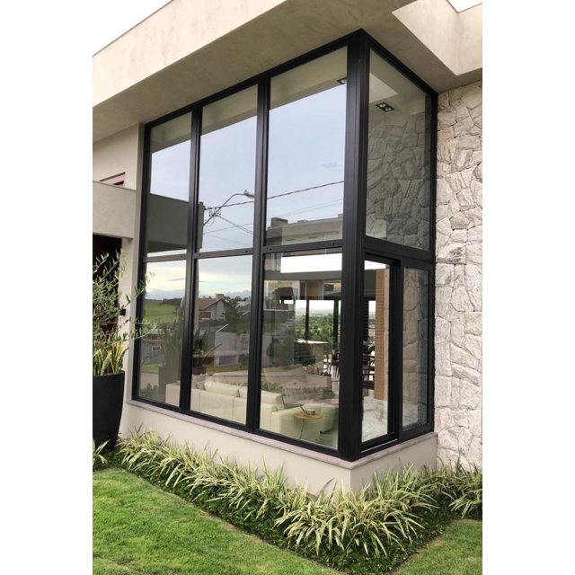 Janelas Fixas e Pele de Vidro para Residências de Alto Padrão Grande janela fixa de canto com esquadrias de alumínio preto, formando uma parede de vidro de pé-direito duplo em uma casa moderna com fachada de pedra.