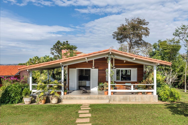 Fachada de uma casa de campo charmosa com paredes de madeira, uma grande varanda com pilares brancos, janelas com venezianas e um amplo jardim gramado.
