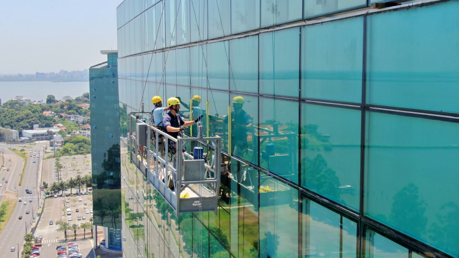 Equipe técnica da Networking Engenharia, com capacetes e equipamentos de segurança, em um andaime suspenso (balancim) realizando a manutenção de uma fachada-cortina.