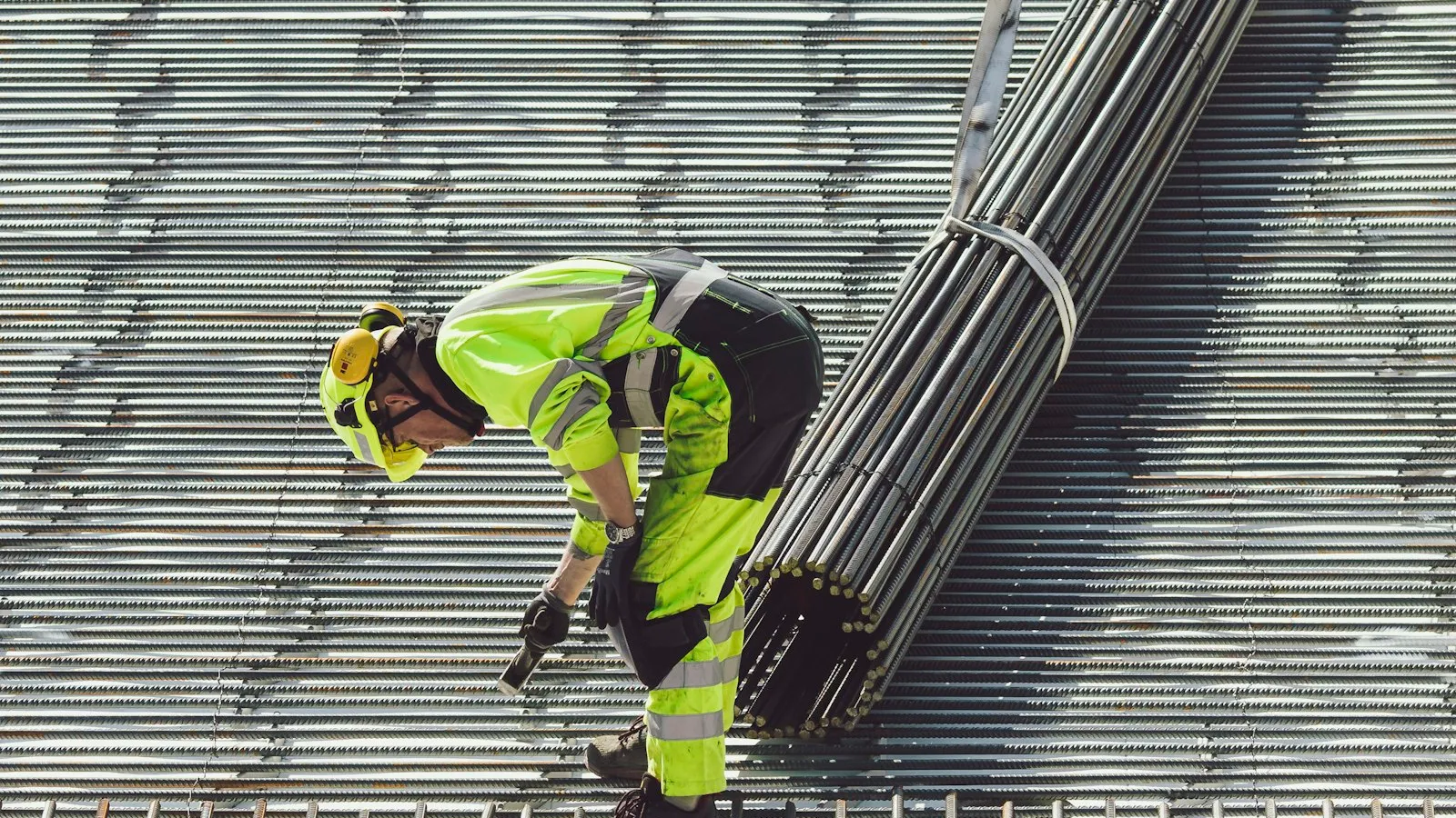 Profissional com equipamentos de proteção manuseando barras de estrutura metálica em obra industrial