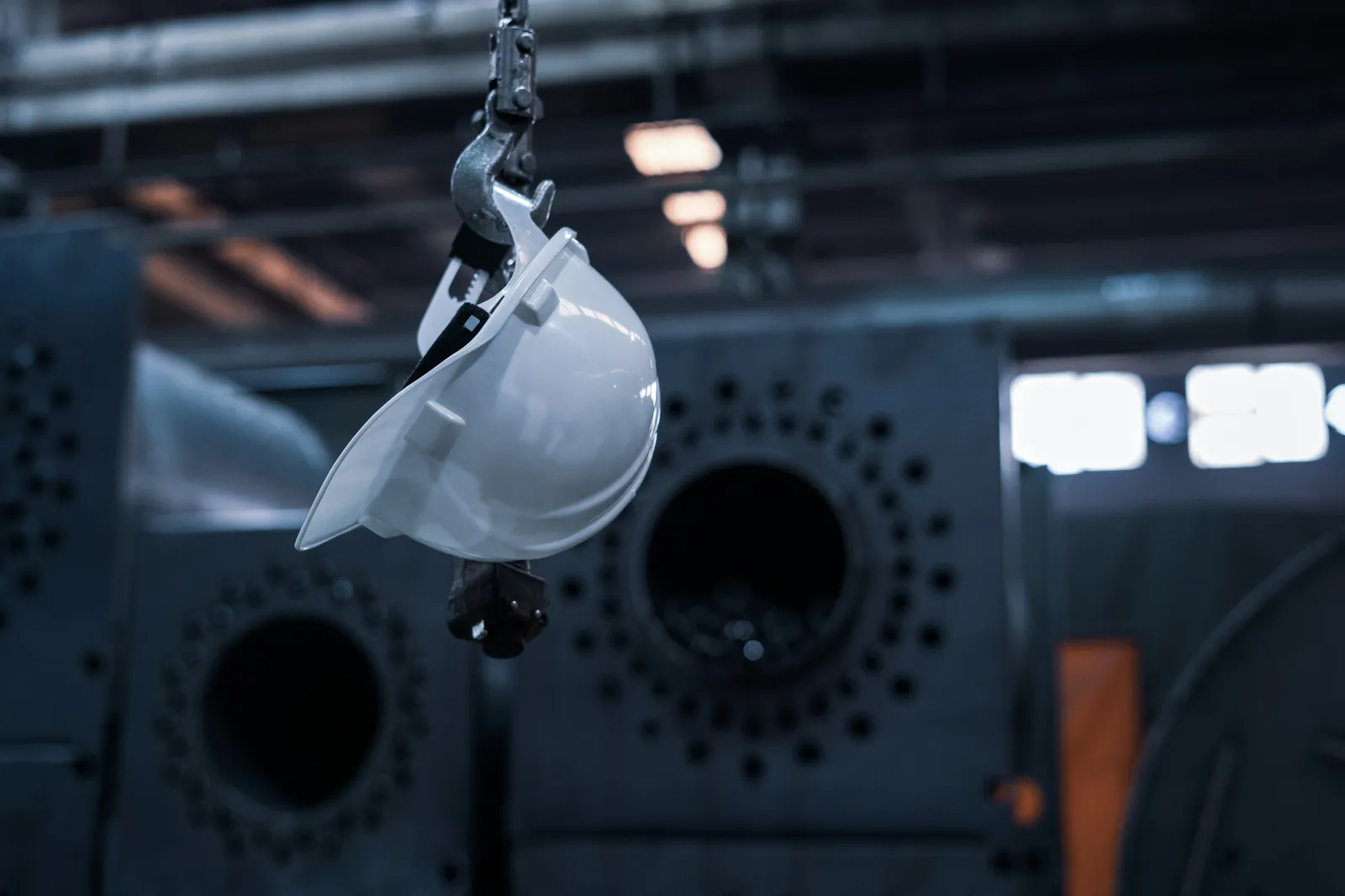 Manutenção predial industrial: equipamentos críticos e segurança do trabalho Capacete de segurança industrial suspenso em gancho de talha dentro de galpão fabril, representando os equipamentos críticos da manutenção predial industrial