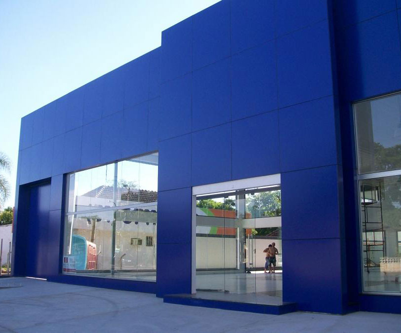 Fachada de loja moderna com revestimento em painéis de ACM azul royal e grandes vitrines e portas de vidro, criando uma entrada convidativa.