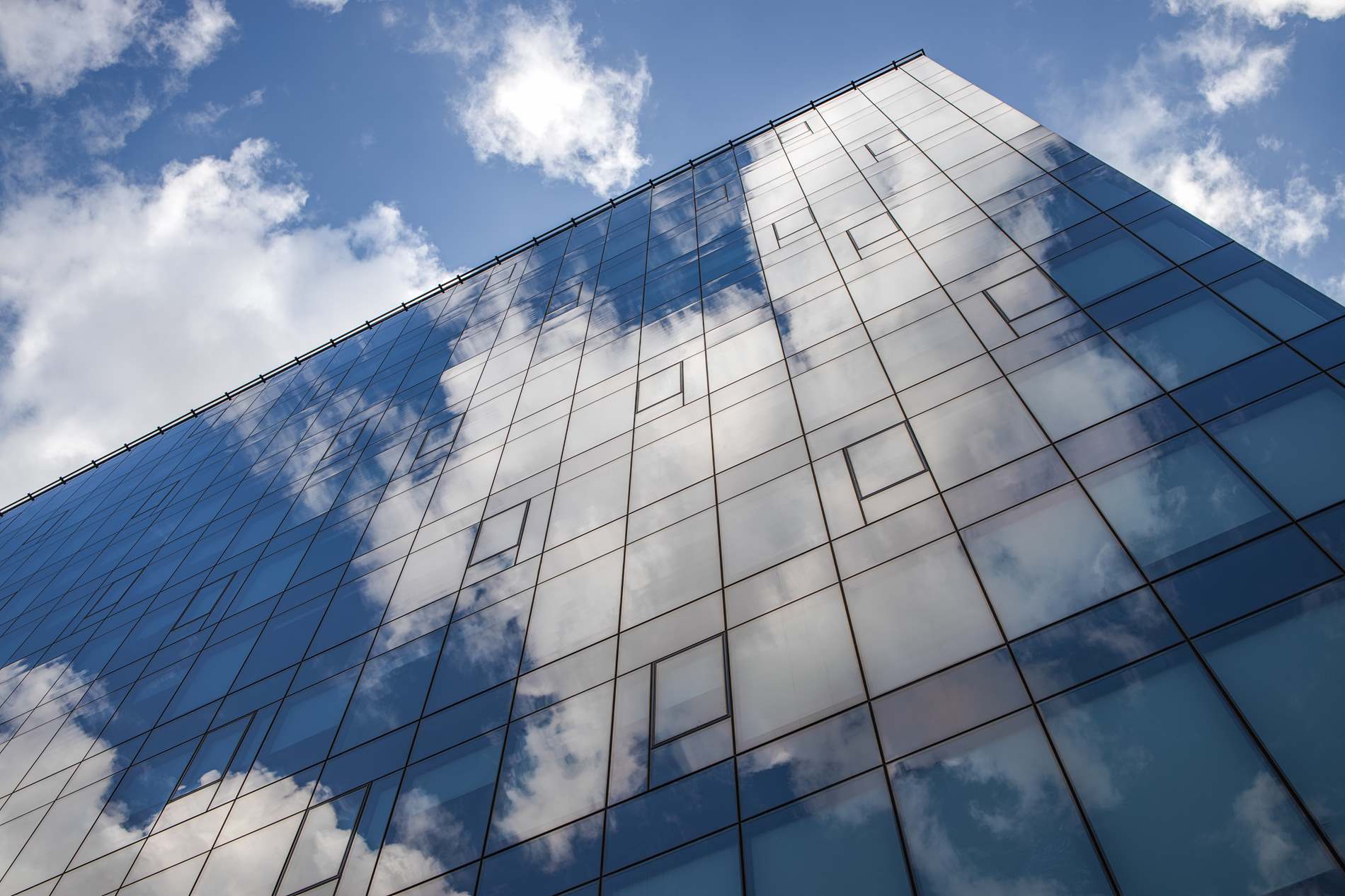 Fachada de um grande edifício comercial com vidro refletivo, espelhando perfeitamente um céu azul com nuvens brancas, ilustrando uma opção moderna para fachadas