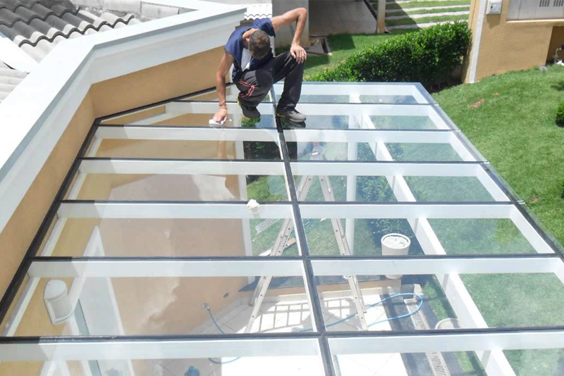 Profissional realizando limpeza e manutenção sobre uma cobertura de vidro com estrutura de alumínio branco instalada em uma varanda residencial.