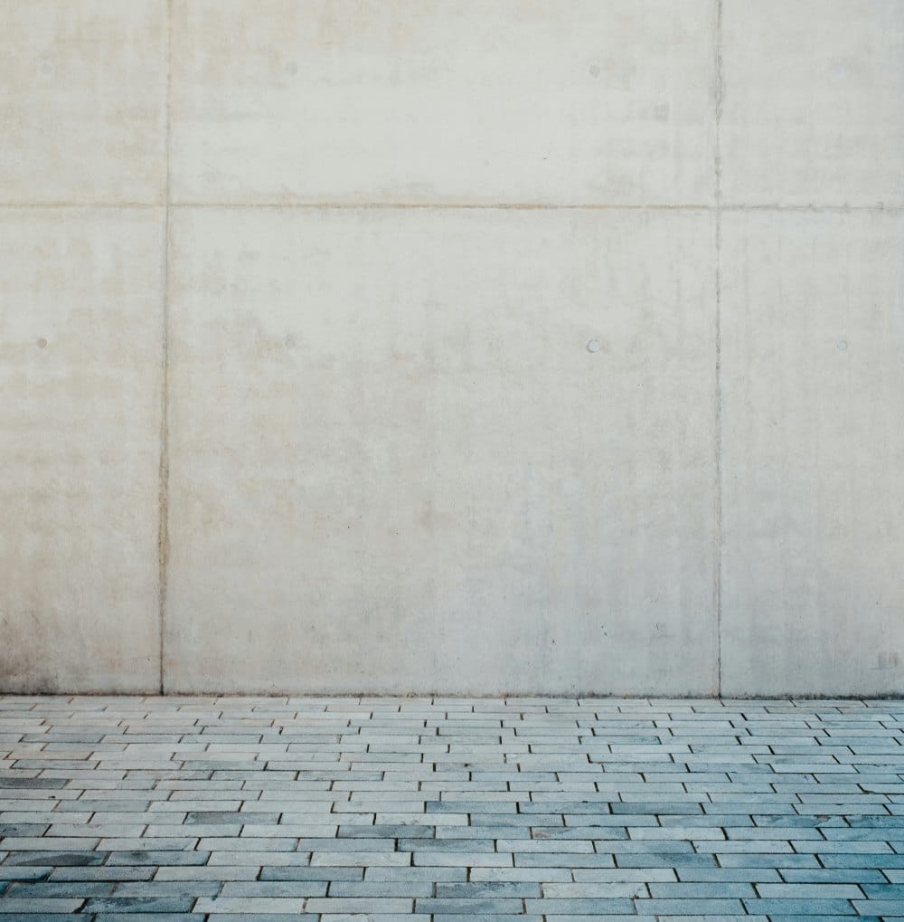 Parede de uma fachada feita com grandes painéis de concreto aparente, com um piso de bloquetes de pedra cinza em primeiro plano.