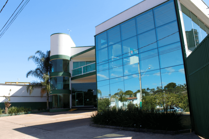 Fachada de um prédio comercial de dois andares com sistema pele de vidro refletivo e uma torre de entrada cilíndrica com detalhes em verde.