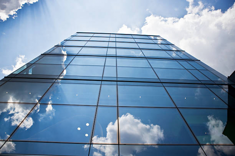 Guia de Preços: Fatores que Influenciam o Custo do Vidro para sua Obra Vista em contraponto da fachada espelhada de um arranha-céu com pele de vidro, refletindo o céu azul e nuvens brancas