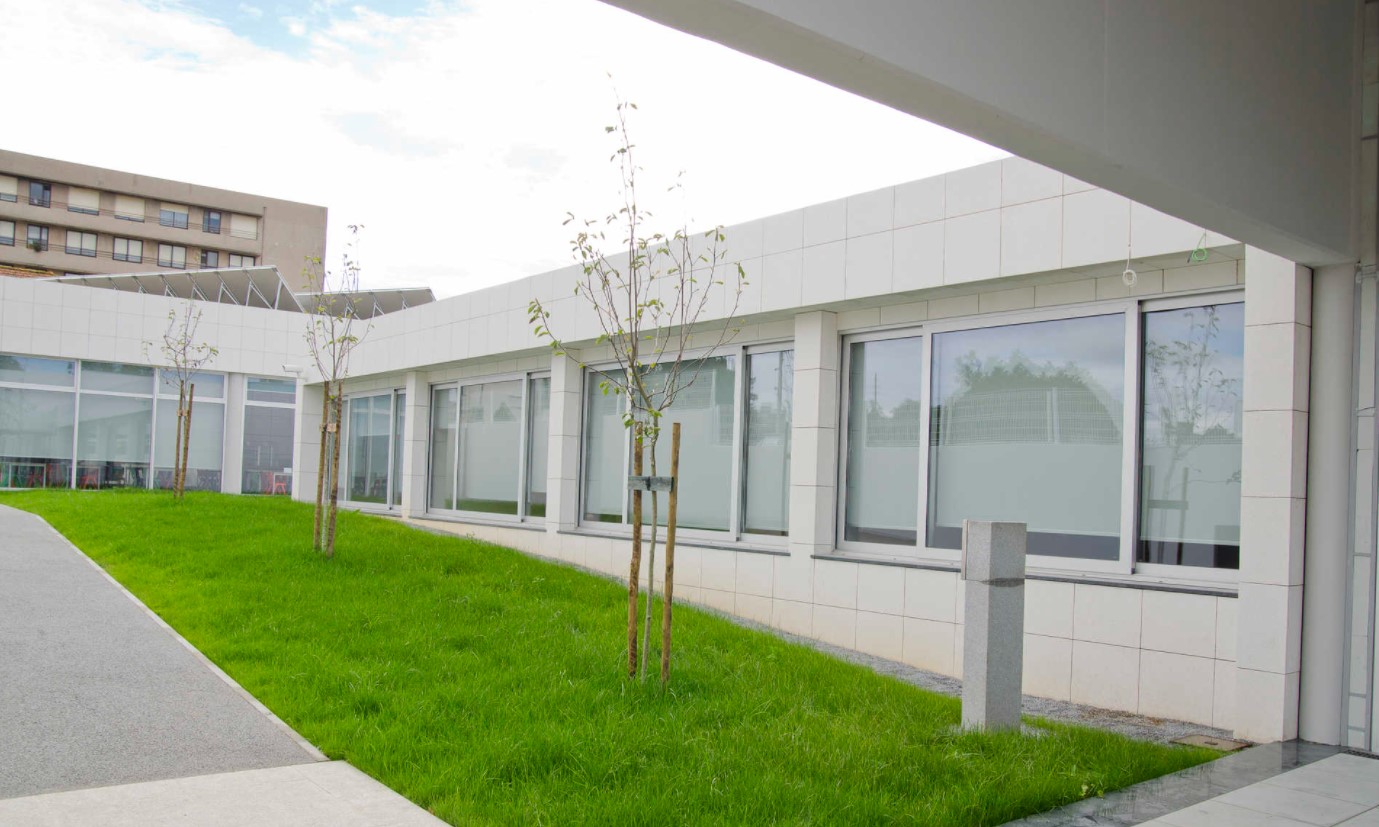 Fachada Escolar com Revestimento de Bloco Vazado: Estética e Funcionalidade A imagem apresenta um exemplo notável de arquitetura escolar contemporânea, onde o revestimento da fachada desempenha um papel central. O andar superior é envolvido por uma pele de blocos vazados (também conhecidos como cobogós), criando um padrão geométrico texturizado que filtra a luz solar e permite a ventilação natural, ao mesmo tempo que oferece privacidade. O térreo contrasta com o uso de madeira e amplas áreas envidraçadas, proporcionando um aspecto acolhedor e transparente que se integra ao pátio gramado. A estrutura de concreto aparente e a passarela metálica complementam o design moderno e funcional do edifício. Este projeto ilustra como a escolha criativa de materiais de revestimento pode valorizar a estética e o conforto ambiental em uma escola.