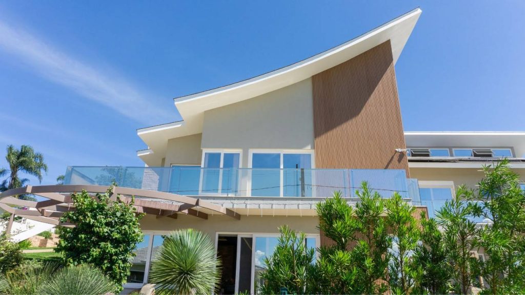 Fachada de uma casa moderna com telhado de abas curvas, revestimento amadeirado (ripado), ampla varanda com guarda-corpo de vidro e um jardim com paisagismo em primeiro plano.