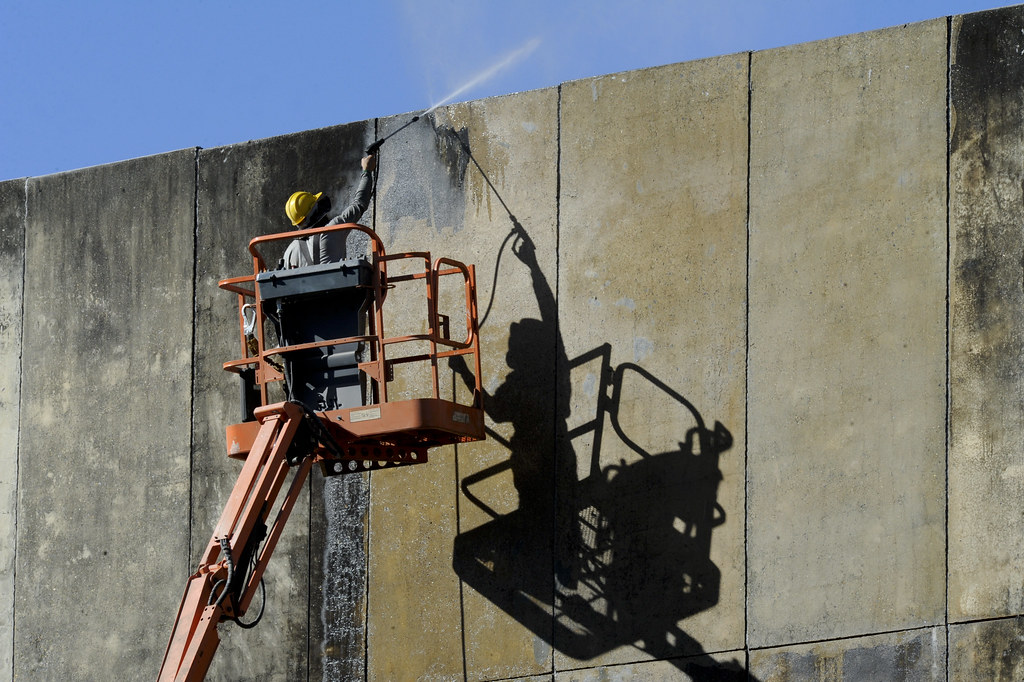 Trabalhador em uma plataforma elevatória (cesta aérea) realizando a limpeza com jato de alta pressão na parede de concreto de uma grande fachada.