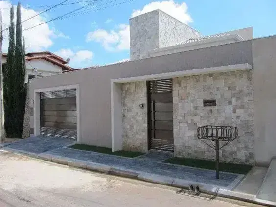 Tipos de Fachada: Um Guia de Materiais e Técnicas para 2025 Fachada de casa térrea moderna com muro revestido em pedra canjiquinha, portão de garagem e porta social de alumínio com ripas horizontais na cor bronze.