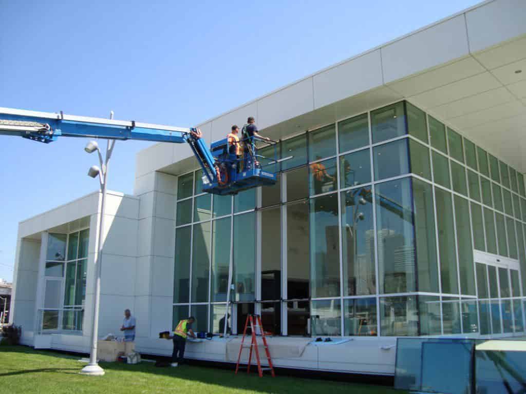 Instalação e manutenção de diferentes tipos de fachadas de vidro em prédios comerciais Equipe técnica realizando a instalação de painéis em uma fachada de vidro de grande porte, utilizando plataforma elevatória articulada para garantir segurança e precisão na obra.