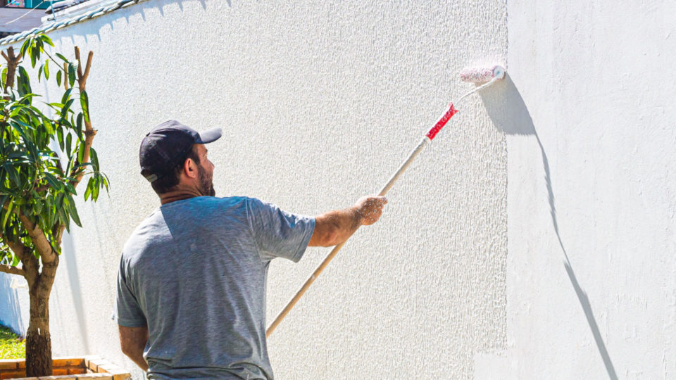 Pintor profissional aplicando tinta branca com rolo de textura em um muro residencial externo, destacando o acabamento e a proteção para superfícies expostas ao sol e chuva.