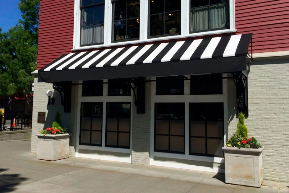 "Fachada de um comércio com um toldo retrátil listrado em preto e branco sobre a porta e as janelas, conferindo um estilo clássico e elegante à loja.