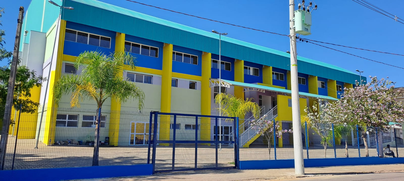Revestimento de Fachada em Cobogó: Ventilação e Conforto Térmico para Escolas Esta imagem exemplifica o uso de blocos de concreto vazados (cobogós) como revestimento principal de uma fachada escolar moderna. No pavimento superior, o material cria uma barreira visual que garante privacidade, enquanto permite a ventilação natural e a entrada de luz filtrada, essenciais para o conforto térmico dos alunos. O contraste entre o concreto, o revestimento em madeira no térreo e as amplas aberturas em vidro confere sofisticação e uma sensação acolhedora ao ambiente educacional, integrando perfeitamente a área externa com o espaço de aprendizagem.