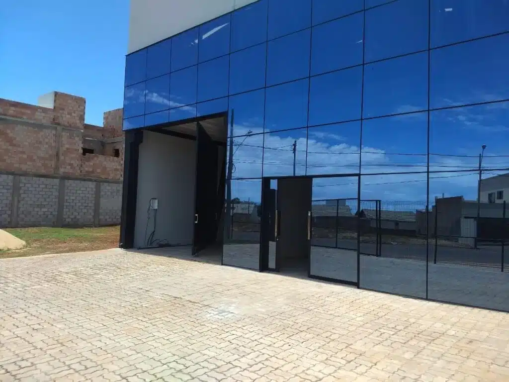 5 Vantagens do Vidro Espelhado para sua Empresa ou Galpão Fachada de um galpão comercial com revestimento completo em pele de vidro espelhado na cor azul, com portas de acesso pretas e piso de bloquetes.