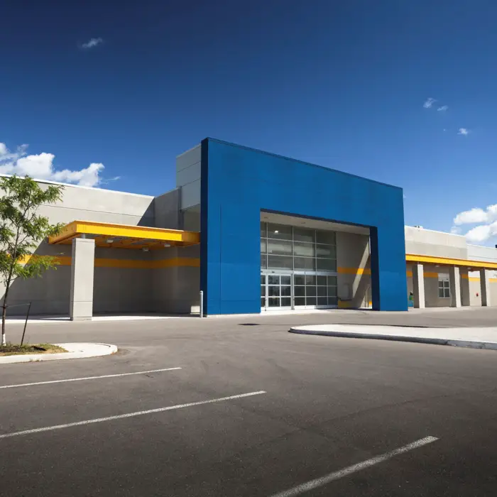 Revestimento em ACM para Casas? Sim! Conheça a Tendência Entrada de um grande edifício comercial ou galpão com fachada moderna, destacando um pórtico revestido em painéis de ACM azul e detalhes em amarelo.