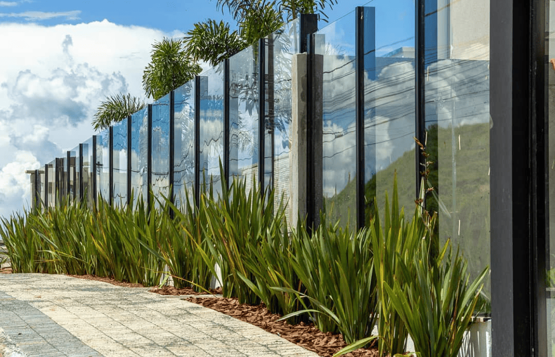 Muro de vidro moderno com painéis espessos de vidro temperado, fixados em robustos postes de alumínio preto, com um jardim de plantas verdes na base.