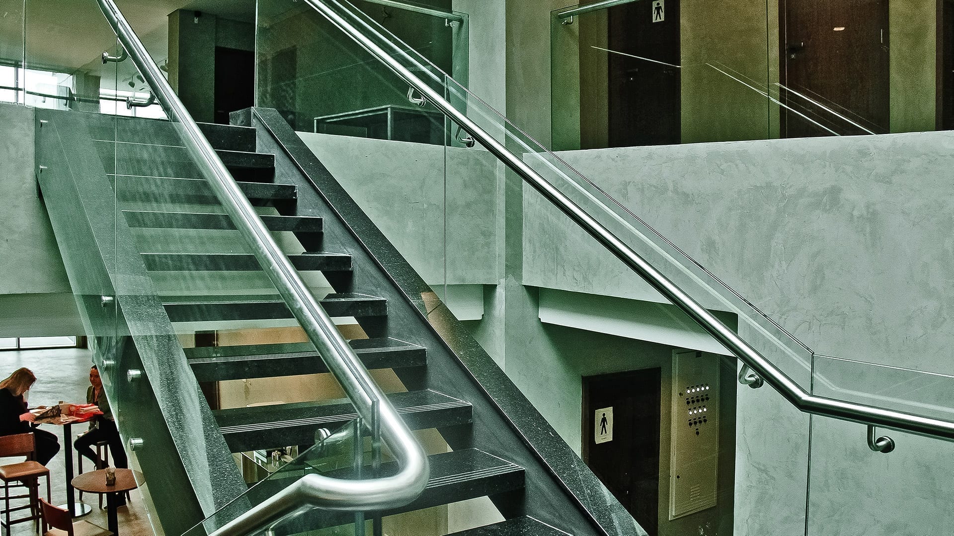 Guarda-Corpo de Vidro: Vantagens e Desvantagens | Vale a Pena? Escada de metal em um ambiente de estilo industrial, com guarda-corpo de vidro e corrimão de aço inox, combinando com as paredes de cimento queimado.