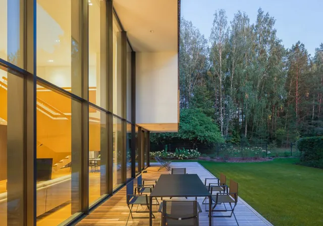 Fachada de uma casa moderna com parede de vidro refletivo na cor champanhe, que se integra a um deck de madeira com vista para um jardim arborizado.