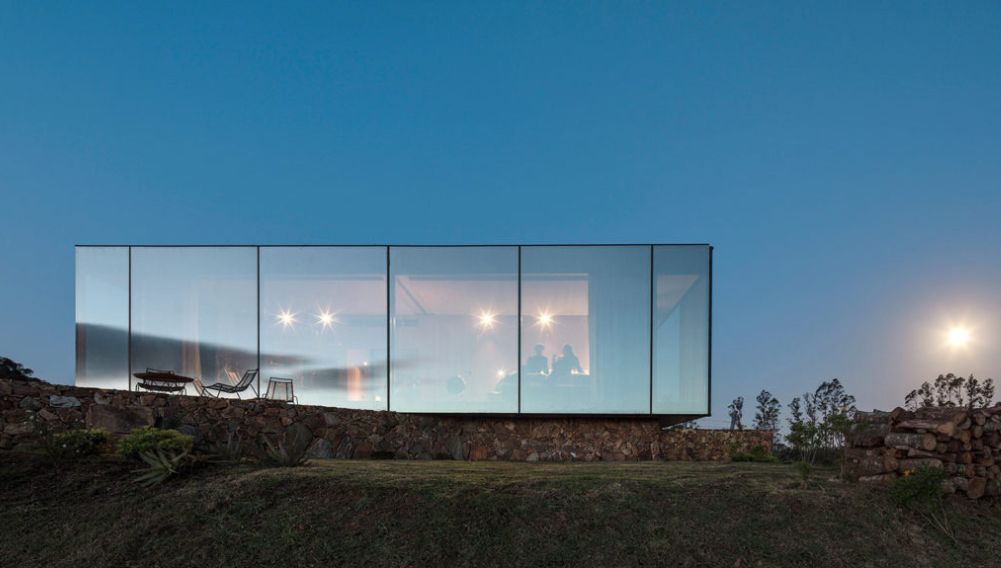 Casa de Vidro do arquiteto Carlos Verna, em Búzios (RJ), ao anoitecer. A estrutura é um cubo de vidro refletivo sobre uma base de pedra, com o interior iluminado.