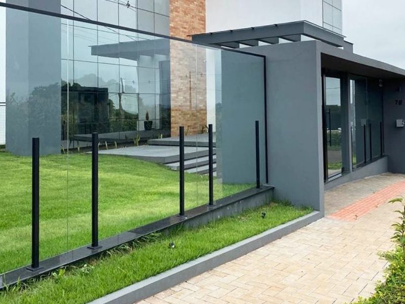 Fachada de casa moderna com muro de vidro temperado sustentado por postes de alumínio preto, ao lado da entrada com paredes de alvenaria cinza.