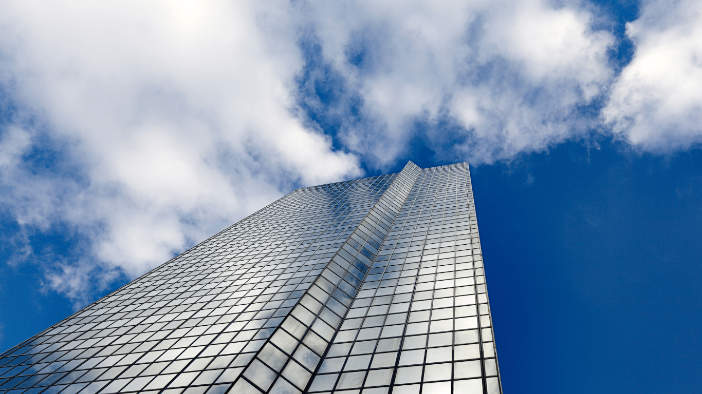 Arranha-céu com fachada de vidro refletindo o céu azul com nuvens, em uma área urbana.